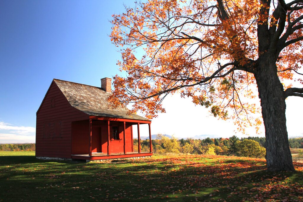 A red farmhouse beside a maple tree in full orange fall color on an open hillside